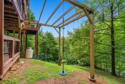 Covered pavilion opening onto the yard and swing, hardwood canopy overhead. Shade, breeze, and a place to sit while the kids burn off the drive.