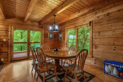 Dining table framed by windows on three sides, hardwood glowing in afternoon light. Cards after dinner, coffee in the morning, forest views the entire time.