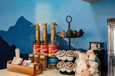 Your morning starts here — a fully stocked coffee bar with Torani syrups, K-cups, teas, and fresh creamers, all set against that signature mountain mural backdrop.
