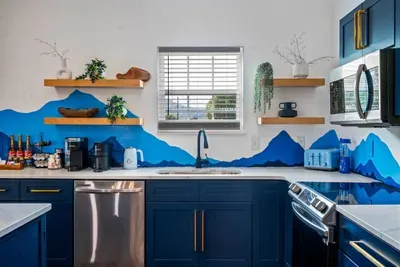 A fully equipped kitchen where the mountain mural follows you in — navy shaker cabinets, quartz counters, stainless appliances, Keurig, Nespresso, and Torani syrups already stocked.