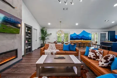 The full sweep of the great room — fireplace lounge, dual leather sofas, a glowing wet bar, and that sweeping mountain mural anchoring every gathering from movie nights to happy hour.