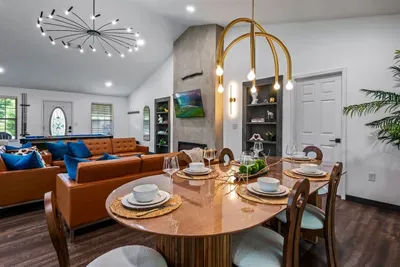 Dinner is served — a marble-topped round dining table set for six, bathed in gold arc chandelier light, with the pool table and great room waiting just steps behind.