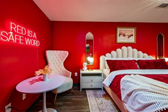 The boldest bedroom in the house — king bed with a sculptural velvet headboard, crimson accent walls, gold mirrors, and a neon sign that sets the whole tone.