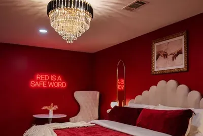 A tiered crystal chandelier crowns the most talked-about bedroom in the house — king bed, crimson velvet, gold arch mirror, and a neon sign that dares you to unwind.