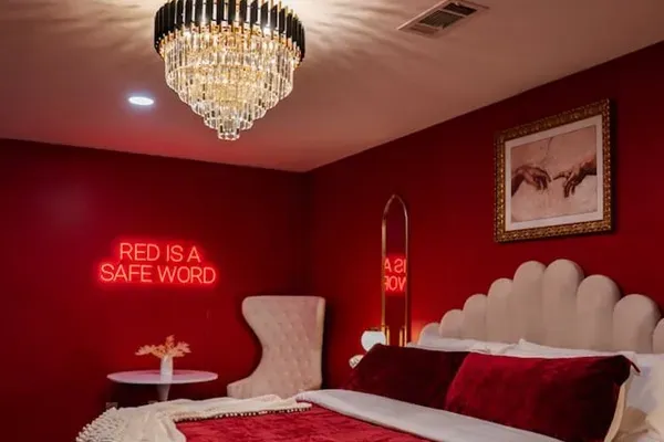 A tiered crystal chandelier crowns the most talked-about bedroom in the house — king bed, crimson velvet, gold arch mirror, and a neon sign that dares you to unwind.