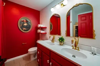 Even the bathroom has range — dual vanity, gold arched mirrors, crimson cabinetry, gold swan faucets, and a sunglasses-wearing Mona Lisa keeping watch over it all.