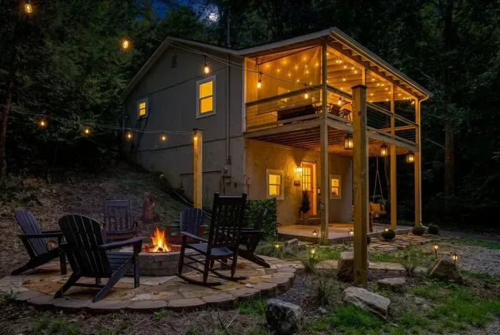 String lights on, fire crackling, cabin glowing through the trees — this is the view of home base every night. Roast marshmallows, count stars, then head up to the deck.