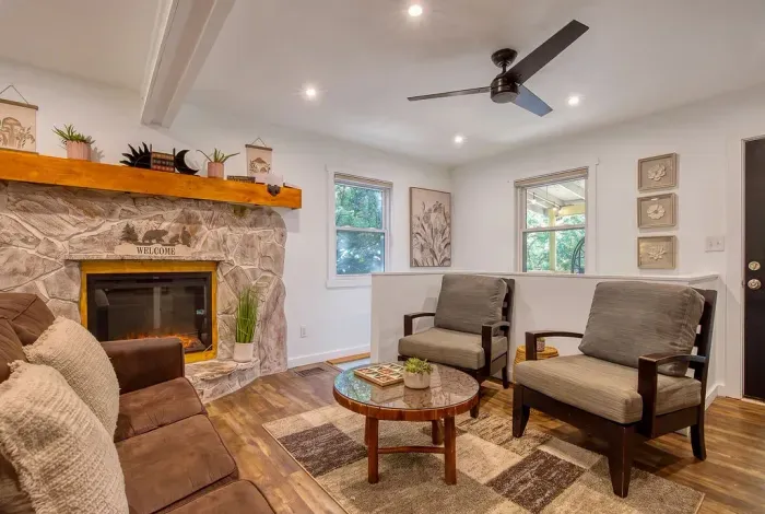 Stone fireplace, plenty of seating, and windows full of trees on both sides. Everyone gets a spot — the sectional, the armchairs, or the floor by the fire with a blanket.