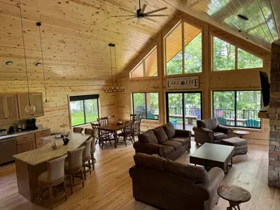 Main level entry view across Lakeside #1 great room, 6-person dining kitchen island, 6-person dining table, family room around gas fireplace, 65" smart TV, access to lake side wrap around deck with BBQ