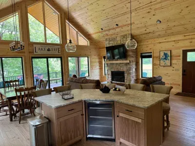 View standing in the middle of the kitchen area looking across the 6-person seating kitchen island with extra beverage refrigerator and outward to the main level living area with gas fireplace, extending out to lakeside wrap around deck