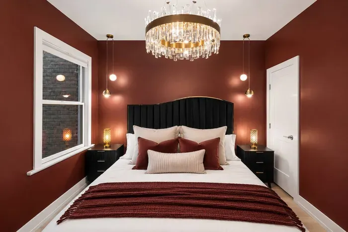 Luxurious king bedroom with rich burgundy walls, a plush black headboard, a statement crystal chandelier, and 55" Roku TV. A dramatic yet cozy retreat designed for restful nights and elegant comfort.