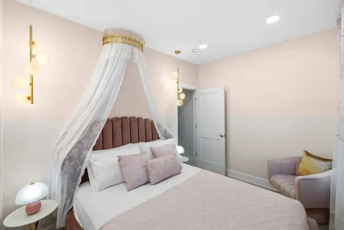 Features a velvet queen-size bed with royal crown canopy, Hollywood vanity mirror, accent chair, bubble pendant lights, and delicate lace curtains. 
