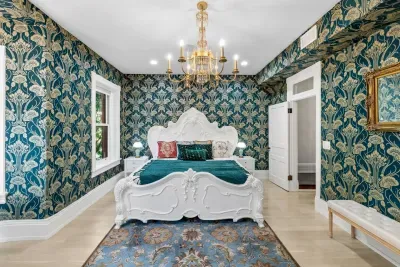 This grand suite features emerald-and-gold wallpaper, two ornate chandeliers, hand-carved white Victorian king bed, writing desk, antique gold wall mirror, full dressing mirror, fireplace, 55” Roku TV, and curated bookshelf of antique volumes.