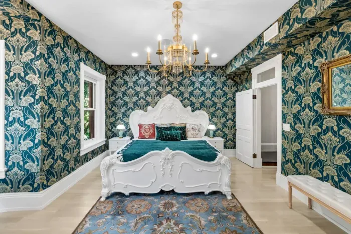 This grand suite features emerald-and-gold wallpaper, two ornate chandeliers, hand-carved white Victorian king bed, writing desk, antique gold wall mirror, full dressing mirror, fireplace, 55” Roku TV, and curated bookshelf of antique volumes. 