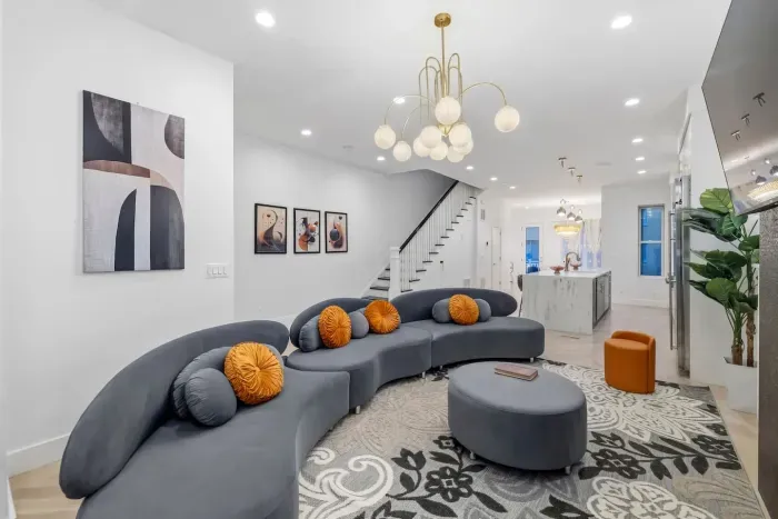 Brown-tiled fireplace, curved velvet sectional with ottoman, chandelier, wall sconce lights, 55'' Roku smart TV, ornate lace drapery, abstract gallery wall, gold bar cart, floor lamp and vase decors. 