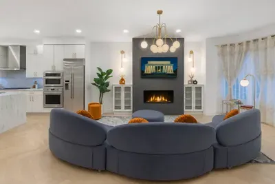 Bright, elegant living room with a curved velvet sofa, warm fireplace, designer chandelier, and bay window view. The space blends historic D.C. charm with modern comfort, creating a stylish gathering area for relaxing, dining, or entertaining.