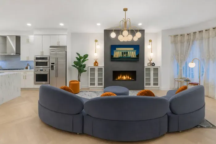 Bright, elegant living room with a curved velvet sofa, warm fireplace, designer chandelier, and bay window view. The space blends historic D.C. charm with modern comfort, creating a stylish gathering area for relaxing, dining, or entertaining.