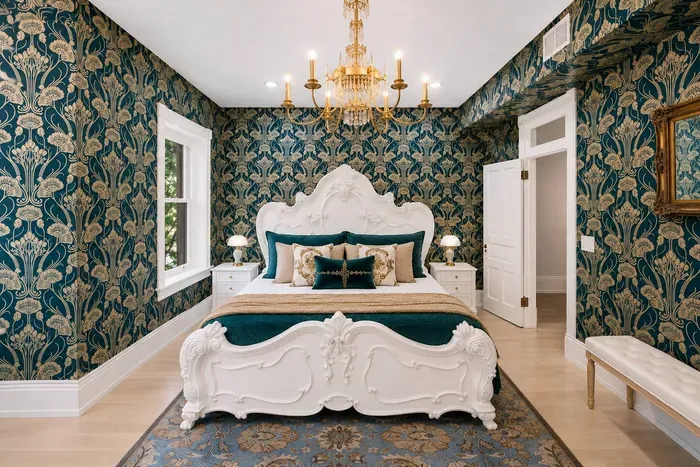 This grand suite features high-end emerald-and-gold wallpaper, two ornate chandeliers, hand-carved white Victorian king bed, work desk, antique gold wall mirror, full dressing mirror, fireplace, 55” Roku TV, and curated bookshelf of antique volumes.