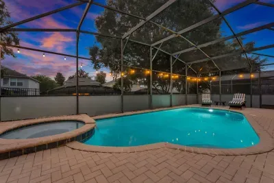 Screened Pool: Freeform pool and adjacent spa glow beneath a vibrant sunset sky, framed by string lights!