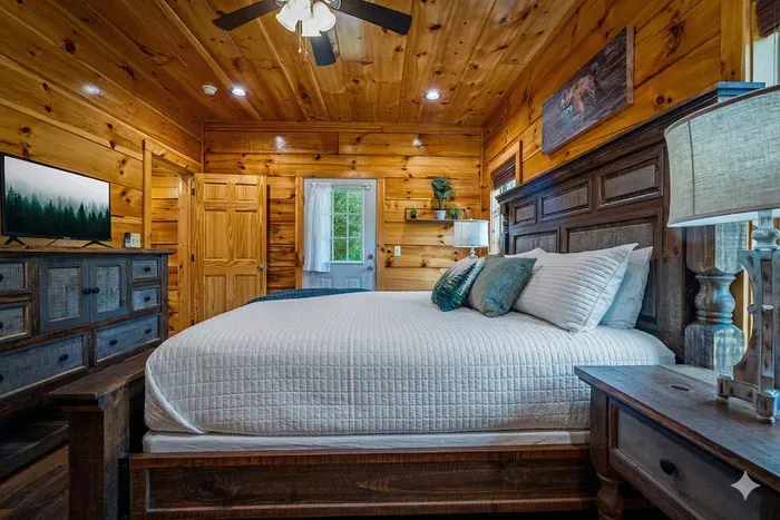 Rustic cabin bedroom with king bed, TV, soft lighting, and ceiling fan