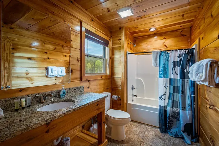 Rustic cabin bath: granite vanity, bright window, tub/shower, stocked linens.