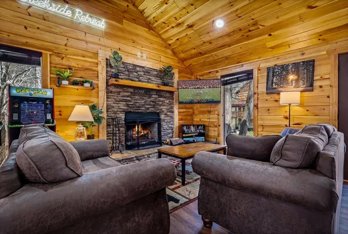Cozy cabin lounge with fireplace, Pac‑Man arcade, TV, and wooded views.