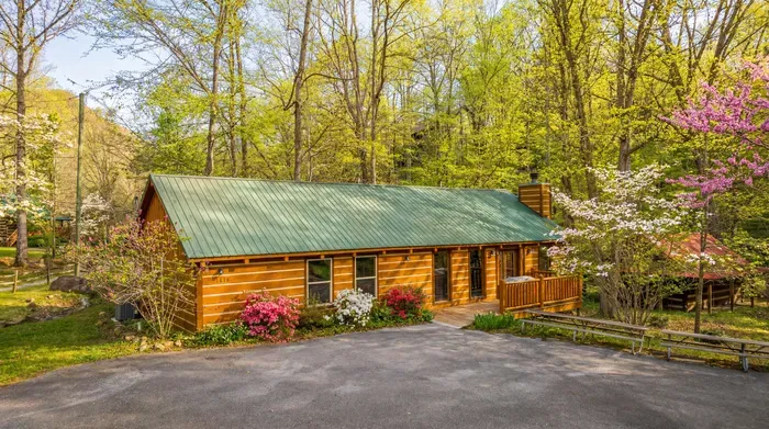 Charming log cabin in the woods with porch, blooms, and paved parking.