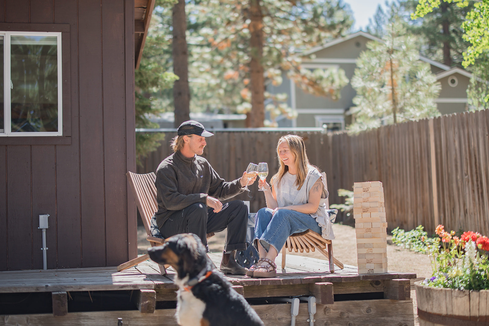Black and tan dog in the foreground, couple with wine glass clinking glasses on a deck with a Jenga set next to them