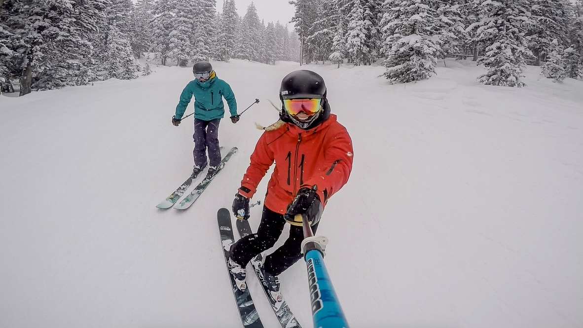 two skiers in a snowy ski slope, one in front with a red jacket and one behind in a teal jacket