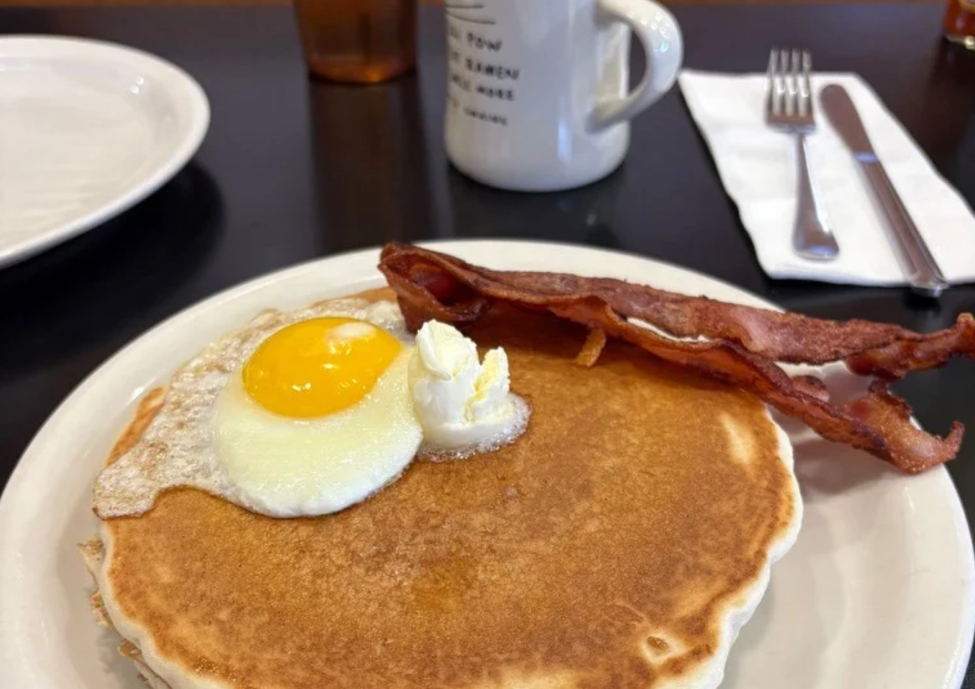 Close up photo of pancakes, bacon, and egg with coffee cup and utensils in the background