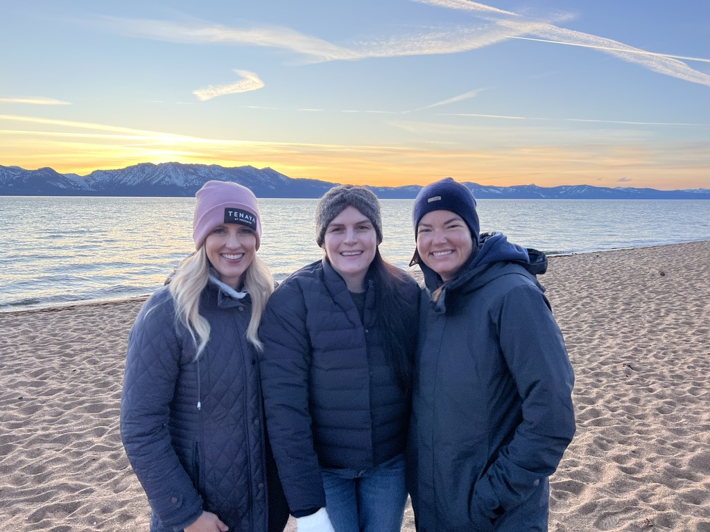 Three women in winter jackets on the shoreline of Nevada Beach in Lake Tahoe, sunset behind them