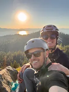 Two people smiling on a mountain bike ride with a lake in the background.