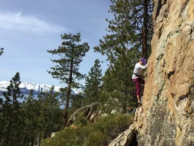 A person rock climing with a white t shirt and purple pants and snow capped mountains in the background.