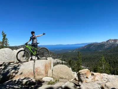 A woman mountainbiking standing on top of boulder looking out at Lake Tahoe.