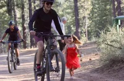 A woman mountain biking with a black shirt on and a little kid running in a red dress next to her.