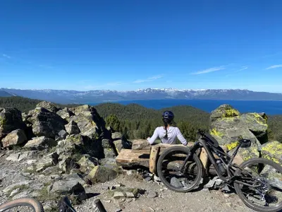 A woman sitting on a bench with a mountain bike leaned up next to it looking out at Lake Tahoe.