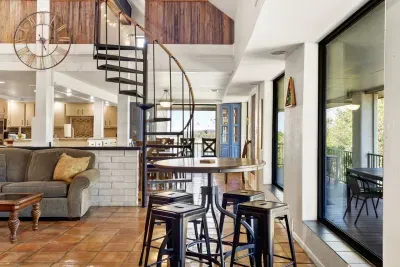 The game area features a cozy 4-seater round table next to the billiards table, with the steel spiral staircase nearby. Beyond, the dining area and kitchen flow seamlessly, all framed by wide windows showcasing beautiful lake views.