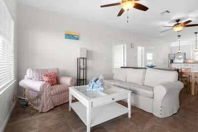 A cozy living space with a striped armchair, a white sofa, and a matching coffee table. The open layout connects to a modern kitchen with stainless steel appliances, wooden furniture, and warm ceiling fans, creating a welcoming and airy atmosphere.