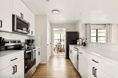 Bright open-concept kitchen and living area with sleek white cabinetry, stainless steel appliances, and wood-style floors. Large windows fill the space with natural light, creating a warm and inviting spot to cook, dine, and relax together.