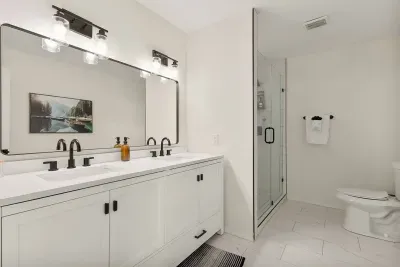 Modern and elegant, this cream-white bathroom features dual sinks, a sleek black-accented faucet, a toilet, and a spacious shower. Perfect for getting ready together or enjoying a relaxing, stylish space.