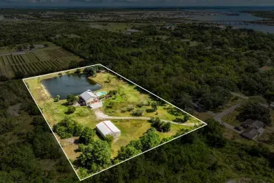 An expansive aerial view of the property and its surroundings. The home, pond, and outbuildings sit within the large, private acreage. Enjoy the mix of open fields and wooded areas, with the bay and city visible on the horizon.