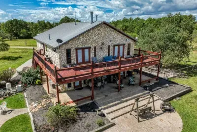 A closer aerial look at the home's stunning stone façade, a massive two-story deck, and the covered patio below. The upstairs features two sets of French doors accessing the deck, providing great views of the large yard and wooded area.