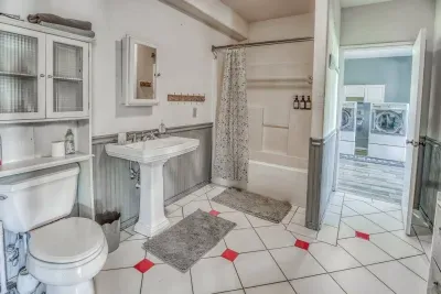 Our clean second bathroom features a tub/shower combo, pedestal sink, and charming wainscoting. Unique white tile with red accents brightens the floor. It conveniently connects to the laundry area with full-sized machines.