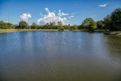 A beautiful, wide view of the tranquil stocked pond on the 5-acre property. Perfect for fishing or simply enjoying the peaceful, private atmosphere and surrounding Texas scenery under a vast blue sky.