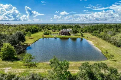 A stunning, wide-angle aerial view of the entire 5-acre property. It highlights the huge stocked pond, the stone house with its private pool, and the peaceful, wooded surroundings. A true private Texas retreat with bay views in the distance.