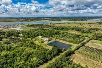 An expansive aerial view of the property and its surroundings. The home, pond, and outbuildings sit within the large, private acreage. Enjoy the mix of open fields and wooded areas, with the bay and city visible on the horizon.