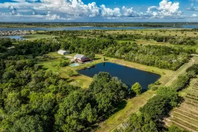 A stunning, wide-angle aerial view of the entire 5-acre property. It highlights the huge stocked pond, the stone house with its private pool, and the peaceful, wooded surroundings. A true private Texas retreat with bay views in the distance.