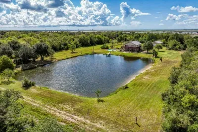 A stunning, wide-angle aerial view of the entire 5-acre property. It highlights the huge stocked pond, the stone house with its private pool, and the peaceful, wooded surroundings. A true private Texas retreat with bay views in the distance.