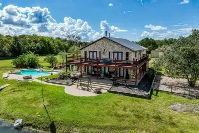 An amazing aerial view of the Two Waters Retreat grounds! The home, with its stone facade and large two-story deck, overlooks the expansive lawn, private swimming pool, fire pit, and a charming wooden bridge leading over the pond inlet.