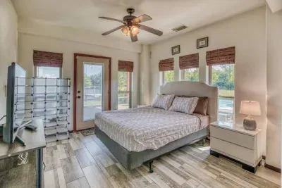Enjoy the bright second bedroom, featuring a Queen bed, wood-look tile floors, and a Smart TV. A glass door provides direct outdoor access, while multiple windows and a ceiling fan ensure a comfortable stay.